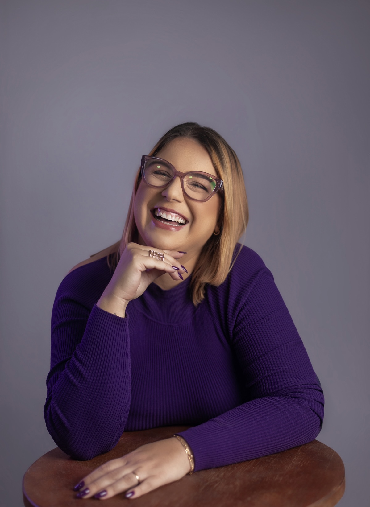 Priscilla Cruz sorrindo, com blusa roxa e a mão sob o queixo